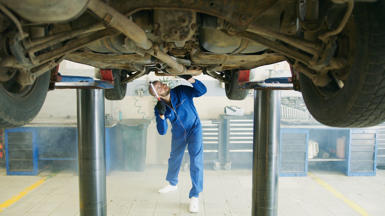 A mechanic in blue uniform inspects a car lifted in a garage workshop.